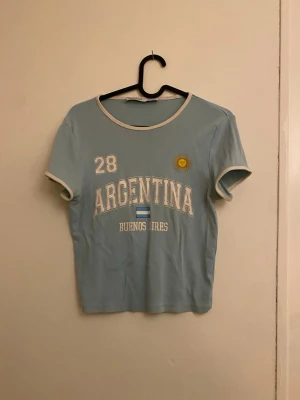 Ljusblå Argentina t-shirt med tryck - Snygg ljusblå t-shirt från Bershka med vit kant vid hals och ärmar. Framsidan har tryck med texten 'Argentina Buenos Aires', siffran 28, en liten sol och flagga. Perfekt för dig som gillar sportig och casual stil.