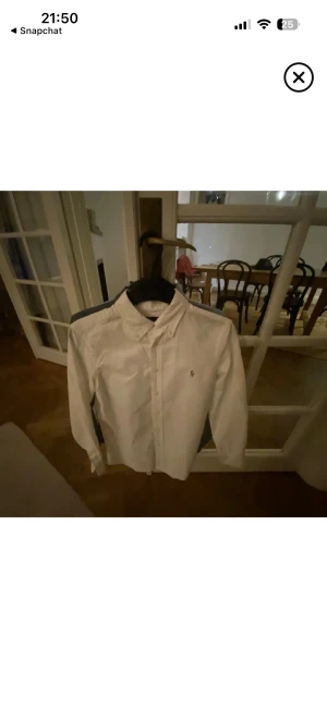 Vit skjorta från Ralph Lauren - Klassisk vit skjorta från Ralph Lauren med broderad logga på bröstet. Skjortan har lång ärm, knappar framtill och en snygg krage. Perfekt för en clean och stilren look.