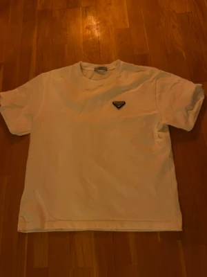 Vit t-shirt från Prada med logga - Vit t-shirt från Prada i mjuk bomull med klassisk rund hals och korta ärmar. På bröstet sitter en svart, triangelformad Prada-logga i metall. Enkel och clean design som ger en lyxig vibe till din outfit.