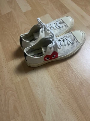 Converse x CDG Play vita sneakers - Säljer ett par vita Converse x Comme des Garçons Play sneakers med det ikoniska röda hjärta-trycket på sidan. Klassisk låg modell med snörning, rund tå och platt sula. Ovandel i slitstark bomullscanvas och gummidetaljer på tån. Perfekt för dig som gillar streetstyle.