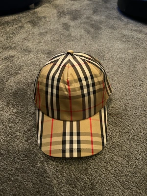  Burberry  - Snygg Burberry keps med klassiskt rutigt mönster i beige, svart, vitt och röda detaljer. Kepsen har böjd skärm och justerbar rem baktill. Materialet är bomull och polyester, vilket gör den både bekväm och stilren. Perfekt för dig som vill ha en ikonisk accessoar.