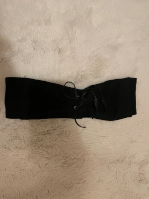 Svart omlottväst med snörning - Säljer en cool svart omlottväst med snörning framtill. Västen är bred och sitter tight runt midjan, perfekt för att ge din outfit en edgy touch. Materialet känns mjukt och har en matt finish. Snörningen gör att du kan justera passformen precis som du vill.