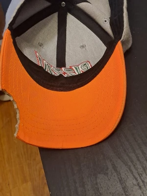 Grå pizza-keps med broderi - Unik keps med grå krona och broderad text 'PIZZA!' i grönt och rött. Skärmen har ett färgglatt pizzamotiv med orange och beige botten samt röda pepperoniskivor. Skärmen har en bit som ser ut att vara 'tuggad' för extra cool detalj.