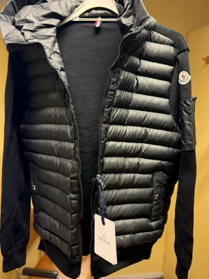 Svart cardigan med luva från Moncler - Svart cardigan med luva från Moncler med quiltad front och ärmar i stickad bomull. Jackan har dragkedja, fickor med tryckknappar och Moncler-logga på ärmen. Snygg och sportig look med blandning av material och klassisk passform.