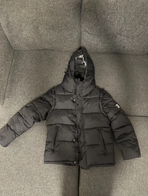 Svart pufferjacka Burberry - Svart pufferjacka Burberry med huva och stora vita logotypdetaljer på ärmen. Jackan har dragkedja och knappar framtill, två sidofickor med dragkedja och quiltad design. Insidan har klassiska Burberry-rutor vid kragen. Perfekt för kalla dagar.den blir även Från winter jacka till Väst