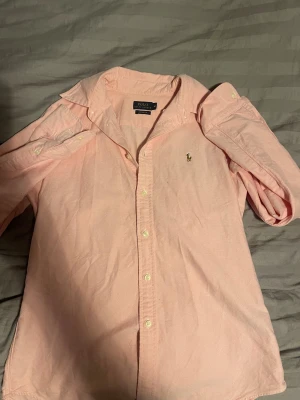 Ljusrosa skjorta från Polo Ralph Lauren - Säljer en ljusrosa skjorta från Polo Ralph Lauren i custom fit. Skjortan har klassisk krage, långa ärmar och knäppning framtill. Broderad logga på bröstet och tillverkad i mjuk bomull. Perfekt för dig som gillar stilrena och snygga plagg. Skjortan är i storlek M 