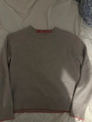 Beige stickad tröja Zadig & Voltaire - Beige stickad tröja från Zadig & Voltaire med rund halsringning och röd texten 'LOVE YOURSELF' på insidan av kragen. Tröjan har ribbade muddar och nederkant samt ett diskret hålmönster vid höften. Perfekt för lager-på-lager och chill vibes.