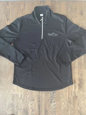 Svart golftröja från Next Tee Golf Clothes - Svart långärmad tröja från Next Tee med vit dragkedja och tryckt logga på bröstet. Perfekt för golf eller chill. Snygg och sportig design med ståkrage och clean look.