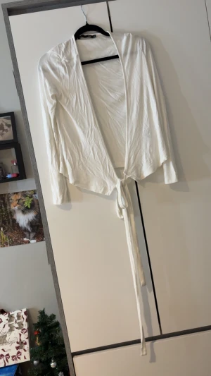 Vit omlottkofta från 157 XS - Snygg vit omlottkofta från 157 i storlek XS. Koftan har långa ärmar och knyts framtill med ett band för en chill och trendig look. Materialet känns mjukt och lätt, perfekt att slänga på över en topp. Passar dig som gillar enkel och stilren design.