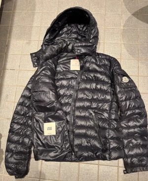Svart Moncler dunjacka med huva - Svart lättvikt dunjacka från Moncler med huva och quiltad design. Jackan har dragkedja framtill, två sidofickor med dragkedja och Moncler-logga på ärmen. Snygg och stilren look med klassisk Moncler-vibe. Det är lagat med tyglim på ett ställe, men om du inte är petig stör det förmodligen inte, och därför är priset satt därefter. Annars är i fint skick, använd inte mycket.  Passar fint storlek M. Från armhåla till armhåla:52 cm. Längd:63 cm
