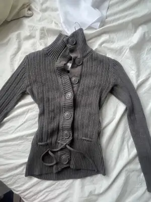 Säljer en grå ribbstickad kofta från H&M L.O.G.G. med hög krage och stora knappar framtill. Koftan har lång ärm, knytband i midjan och är tillverkad i mjuk bomull. Perfekt för dig som gillar chill och stilren look.