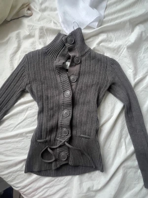 Grå ribbad kofta från H&M L.O.G.G. - Säljer en grå ribbstickad kofta från H&M L.O.G.G. med hög krage och stora knappar framtill. Koftan har lång ärm, knytband i midjan och är tillverkad i mjuk bomull. Perfekt för dig som gillar chill och stilren look.