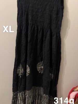 Svart långklänning med mönster XL - Svart långklänning i storlek XL med unika vita mönster och rynkad överdel. Nederdelen har ett randigt mönster i svart och vitt som ger en cool kontrast. Perfekt för dig som gillar bohemisk stil och vill ha något som sticker ut.🌸 Fint använt skick. Storlek XL