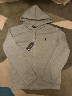 Grå Polo Ralph Lauren hoodie - Grå polo-hoodie i mjukt och bekvämt material. Stilren modell med polokrage som ger ett unikt och trendigt uttryck. Perfekt till både vardag och mys. 