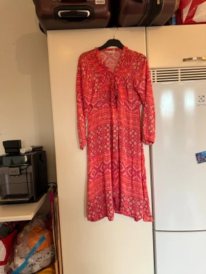Rosa långklänning med blommigt mönster - Snygg långklänning i korallrosa och orange och rosa toner med blommigt och geometriskt mönster. Klänningen har lång ärm, v-ringning med snörning och är gjord i mjuk, tunn viskos. Perfekt för dig som gillar boho-vibe och färg! Från märket Odd Molly. Storlek 2 som enligt deras storleksguide motsvarar en M. Men den är något liten i storlek. Mycket fint skick, jag hittar inga synliga anmärkningar eller defekter. 