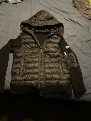 Svart Moncler pufferjacka med huva moncler cardigen  - Svart pufferjacka från Moncler i storlek 160 till 170 med stickade ärmar och huva. Jackan har quiltad front, dragkedja, fickor med knappar och en extra ficka med dragkedja på ärmen. Moncler-logga på vänster ärm och coolt serietryck på insidan. Det går att hålla mobilen mot moncler märket så kommer moncler hemsidan upp