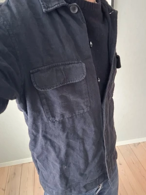 Svart overshirt från Selected Homme - Svart overshirt från Selected Homme med klassisk krage och två stora bröstfickor med lock. Jackan har knäppning framtill och är tillverkad i mjuk bomull. Perfekt att slänga på över en t-shirt eller hoodie för en chill look. Storlek S!