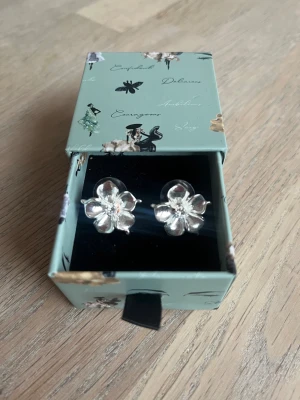 Ioaku La Fleur Örhänge - Säljer ett par snygga örhängen i form av blommor från Ioaku. De har fem kronblad och en liten detalj i mitten som ger extra bling. Perfekta för dig som vill ha något gulligt men ändå stilrent till din outfit.