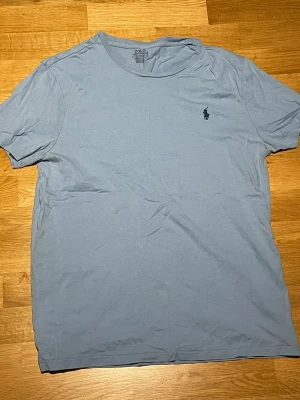 Ljusblå t-shirt från Polo Ralph Lauren - Snygg ljusblå t-shirt från Polo Ralph Lauren med klassisk logga broderad på bröstet. T-shirten har rund hals och korta ärmar, perfekt för en chill och stilren look. Tillverkad i mjuk bomull som känns skön mot huden.