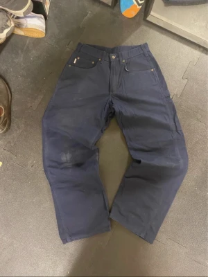 Mörkblå cargopants från Carhartt - Säljer ett par mörkblå cargopants från Carhartt med klassisk logopatch bak. Byxorna har raka ben, flera praktiska fickor och robust bomullstyg. Perfekta för dig som gillar streetwear och vill ha en chill men funktionell look.