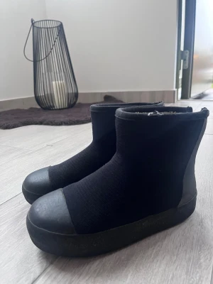 Svarta boots med dragkedja och foder - Säljer ett par svarta boots med rund tå och platt sula. Skorna har dragkedja bak och är fodrade med mjukt material på insidan. Ovandelen är i textil och tå samt sula i gummi, vilket ger en clean och modern look.