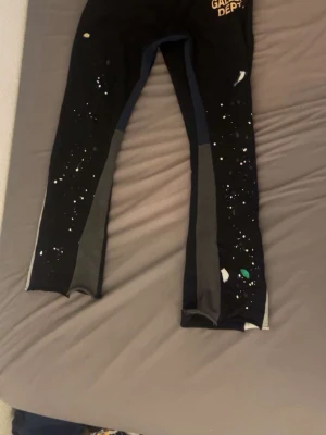 Gallery dept mjukis byxor med galaxmönster - Säljer ett par svarta leggings från Gallery Dept. med coolt galaxinspirerat mönster i vitt och färgdetaljer. Byxorna har kontrasterande grå och blå paneler längs benen och en smal passform som sitter tight. Perfekt för dig som gillar unika och kreativa plagg.