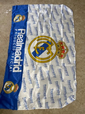 Real Madrid officiell flagga - Stor och snygg Real Madrid flagga med klubbens klassiska logga i blått, gult och rött på vit bakgrund. Flaggan har blå kant med tryckt text och flera små loggor. Perfekt för fotbollsfans som vill visa sitt stöd för laget.
