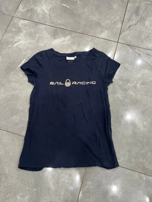 Mörkblå Sail Racing t-shirt XS - Snygg mörkblå t-shirt från Sail Racing i storlek XS. T-shirten har korta ärmar och rund hals, med ett stilrent Sail Racing-tryck i vitt på bröstet. Tillverkad i mjuk bomull som känns skön mot huden. Perfekt för dig som gillar sportig och enkel stil.