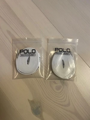 Skosnören | RALPH LAUREN. - Två par vita skosnören från Polo Ralph Lauren, levereras i originalförpackning med logga. Skosnörena är platta och har en design med Polo-tryck längs hela snöret. Perfekt för att ge dina sneakers eller skor en fräsch och clean look.