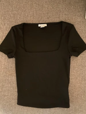 Svart topp med fyrkantig hals från newyorker - Säljer en svart topp från newyorker i storlek XS. Toppen har en snygg fyrkantig halsringning och korta ärmar. Materialet är mjukt och stretchigt, perfekt för att styla med jeans eller kjol. Enkel och stilren design som funkar till många looks.