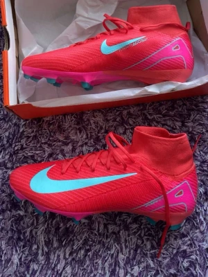 Nike röda fotbollsskor med turkos swoosh - Säljer ett par grymma Nike fotbollsskor i röd färg med turkos swoosh och detaljer. Skorna har snörning, hög krage och platt sula med dobbar i rosa och turkos. Materialet är syntet och skon har en strömlinjeformad design som sticker ut på planen fråga gärna om ni vill se fler bilder.