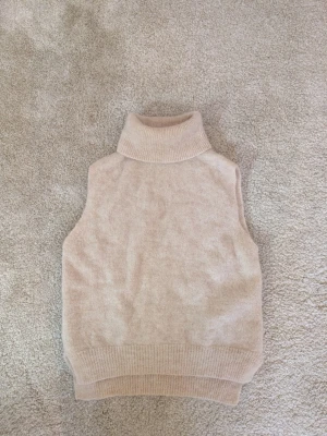 Beige stickad tröja utan ärmar från Bik Bok - Mysig beige stickad väst från Bik Bok i alpackablandning. Modellen har hög polokrage och ribbade kanter nedtill och vid halsen. Perfekt att styla över en långärmad tröja för extra lager. Enkel och stilren design utan mönster. Utan armar