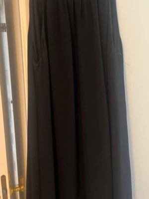 Svart långklänning med pärldetaljer - Svart långklänning med ärmlös design och rundad hals. Halsen är dekorerad med glittrande pärlor och stenar i ett snyggt mönster. Klänningen har ett mjukt, fallande tyg som ger en elegant siluett. Perfekt för dig som gillar stilrena plagg med extra detaljer.