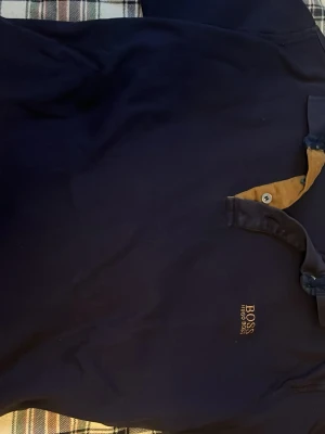 Marinblå polotopp från Hugo Boss - Säljer en stilren marinblå polotopp från Hugo Boss med gul insida på kragen och broderad logga på bröstet. Toppen har klassisk krage med knappar och är perfekt för dig som gillar enkel och snygg design.