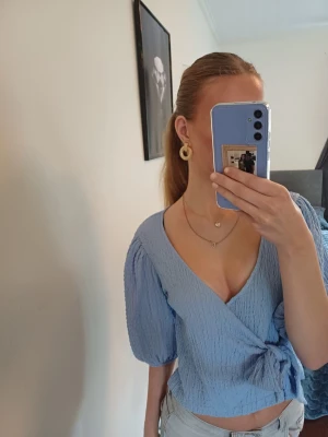 Ljusblå topp/blus med knyt - Supersöt ljusblå ttopp/blus med knyt i sidan och puffiga trekvartsärmar. Toppen har en djup v-ringning. Perfekt för vår och sommar. Storlek M men passar även S