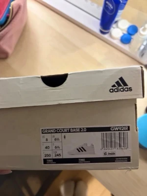 Adidas Grand Court Base 2.0 sneakers - Säljer ett par vita Adidas Grand Court Base 2.0 sneakers med klassiska svarta ränder på sidan. Skorna har en stilren och tidlös design med låg profil och platt sula. Perfekta för dig som gillar en clean look och vill ha bekväma skor till vardagen.