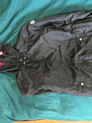 Moncler jacka - Svart vindjacka från Moncler med huva och hel dragkedja framtill. Jackan har diskreta logodetaljer på ärmen och innerficka med Moncler-märke. Materialet är lätt och glansigt polyester, perfekt för blåsiga dagar. Snygg och enkel design med metallknappar på fickorna. Köpt på Tise, pris går absolut att diskutera