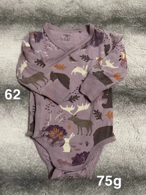 Lila omlottbody med djurmönster från Lindex - Säljer en lila omlottbody från Lindex med lång ärm och tryckknappar. Bodyn har ett gulligt djurmönster med älgar, harar och fåglar i naturfärger som beige, brun och vit. Perfekt för dig som gillar söta och lekfulla prints.🌸 Fint använt skick. Storlek 62