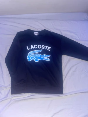 Lacoste tröja - Snygg Lacoste sweatshirt i mörkblå färg med stor ikonisk krokodillogo i blått och vitt framtill. Klassisk rund hals och bekväm passform – perfekt både till vardags och en mer sportig stil.  🔹 Märke: Lacoste 🔹 Färg: Mörkblå/svart 🔹 Skick: Mycket fint skick, (inte använd) 🔹 Modell: Sweatshirt / collegetröja 🔹 Storlek: M passar S