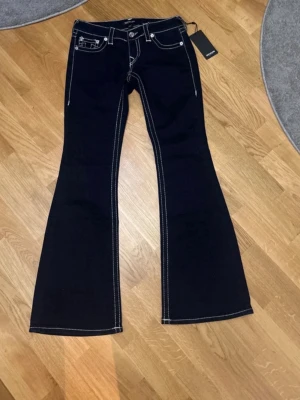 True Religion bootcut jeans, mörk blå - Mörkblå jeans från True Religion med tydliga vita kontrastsömmar och klassiska broderade bakfickor. Modellen är bootcut med låg midja och har metallknappar samt dragkedja. Materialet är jeans och passformen är loose. Perfekt för dig som gillar statement-jeans.