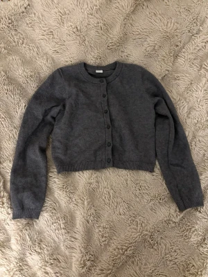 Mörkgrå stickad kofta med knappar - Säljer en mörkgrå, croppad stickad kofta med rund hals och långa ärmar. Koftan har knappar framtill och ribbade muddar vid ärmslut och nederkant. Perfekt att slänga över en t-shirt eller linne för extra värme.