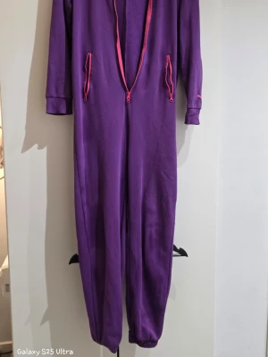 Lila jumpsuit från Puma med rosa detaljer - Säljer en lila jumpsuit från Puma med rosa dragkedjor och logga. Mjuk och skön bomull, långa ärmar och muddar vid benslut och ärmslut. Perfekt för chill dagar eller när du vill sticka ut med färg. Två fickor med dragkedja framtill.