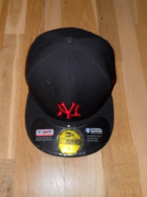 Svart New Era 59FIFTY keps - Säljer en svart New Era 59FIFTY keps med röd broderad NY-logga framtill och MLB-logga baktill. Kepsen har en klassisk rak skärm och är tillverkad i ett slitstarkt material med Cool Base-teknologi för extra komfort. Perfekt för dig som gillar streetwear och sportig stil. Pris kan diskuteras 