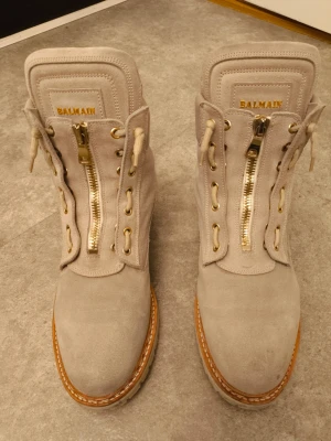 Beige mockaboots från Balmain - Säljer ett par snygga beige boots från Balmain i mjuk mocka med grov sula och rund tå. Skorna har guldfärgade detaljer, snörning och dragkedja framtill samt Balmain-logga på plösen. Perfekt för dig som gillar exklusiva och stilrena boots. Nypriset var ca 15000. Skorna tillverkas inte längre och är därmed unika och i väldigt bra skick. Pris kan diskuteras.