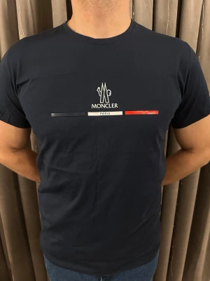 Mörkblå Moncler t-shirt med tryck - Snygg mörkblå t-shirt från Moncler med klassiskt tryck på bröstet i svart, vitt och rött. T-shirten har rund hals och korta ärmar. Tillverkad i mjuk bomull som känns skön mot huden. Perfekt för dig som gillar stilrena och exklusiva plagg.