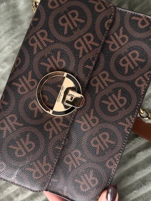 Brun axelväska från Cerruti 1881 - Säljer en snygg brun axelväska från Cerruti 1881 med kedjerem och läderdetalj. Väskan har ett grafiskt mönster med logotypen RR över hela ytan och ett elegant guldfärgat spänne framtill. Perfekt storlek för det viktigaste och riktigt trendig design.