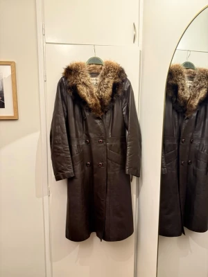 Vintage brun skinnrock med äkta päls   - Säljer en brun lång skinnrock från TerhuKarin, Finland. Rocken har en stor fluffig äkta päls runt kragen i bruna och beige toner, dubbla rader med knappar och två stora fickor framtill. Perfekt för dig som vill sticka ut med vintagevibbar. Liten i storlek, står 38 men mer som 36