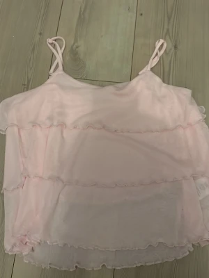 Ljusrosa volang linnetopp - Säljer en ljusrosa linnetopp med tunna axelband och flera lager volanger. Toppen är ärmlös och har en romantisk, flowy känsla som passar perfekt till varma dagar. Materialet känns lätt och luftigt, vilket gör den skön att bära under sommaren.