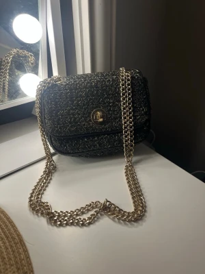 Svart axelväska med guldkedja - Snygg svart axelväska med flätad struktur och inslag av guldtråd. Väskan har en guldfärgad kedja som axelrem och ett vridlås i guld på framsidan. Perfekt storlek för det viktigaste och ger en cool touch till din outfit.