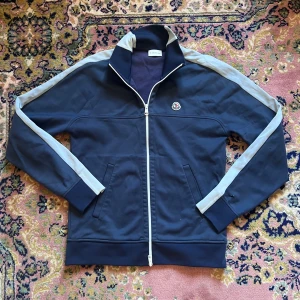 Moncler marinblå Cardigan med ränder - Snygg marinblå ziptröja från Moncler med vita ränder längs ärmarna och broderad logga på bröstet. Tröjan har hög krage, hel dragkedja och två fickor framtill. Perfekt för en sportig och stilren look. Litet bränt hål på höger arm ungefär som ett lillfinger i diameter 